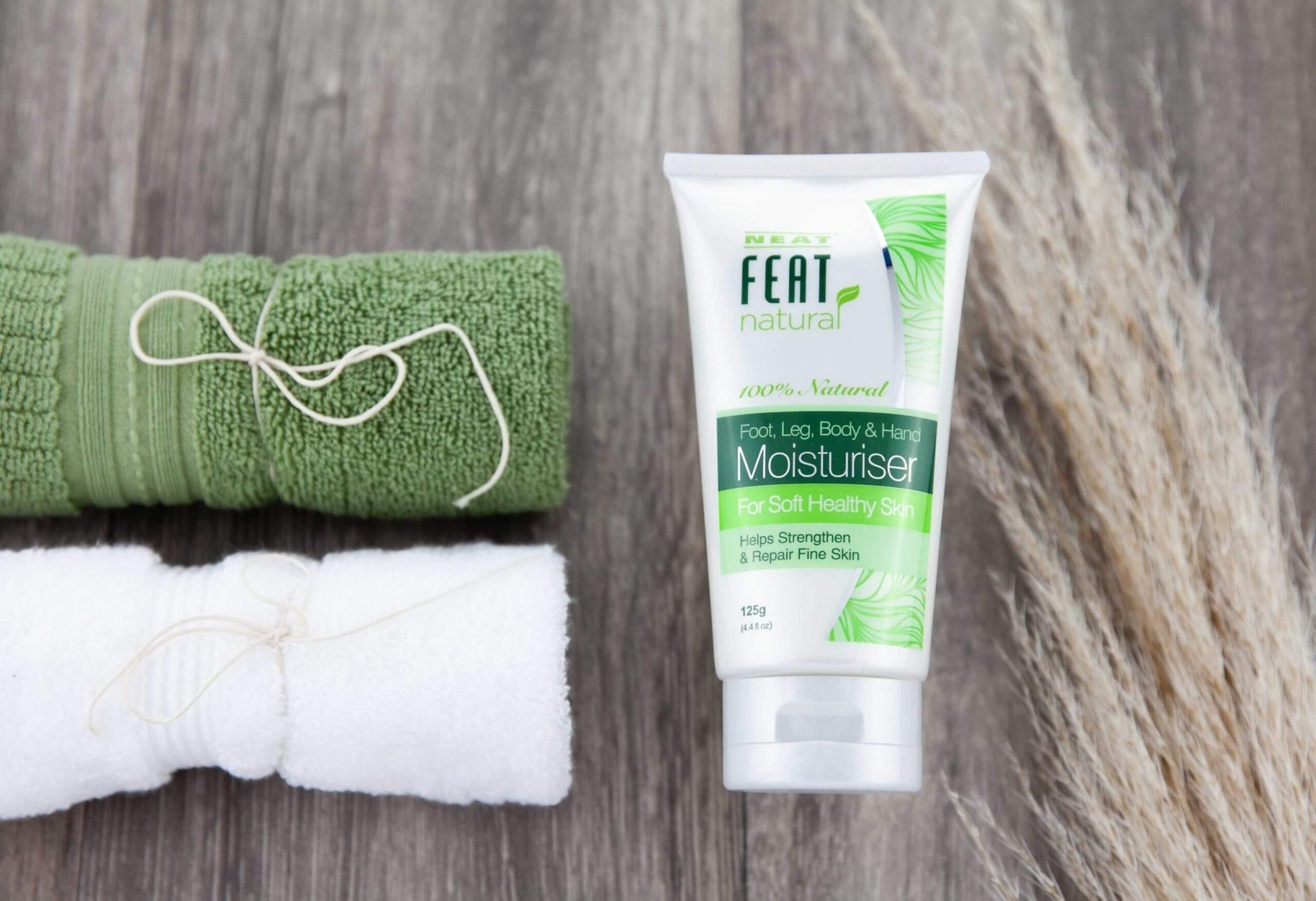 Neat Feat Natural Foot, Leg, Body & Hand Moisturiser tube, laying on a natural-looking table with a green and white small towel beside it.